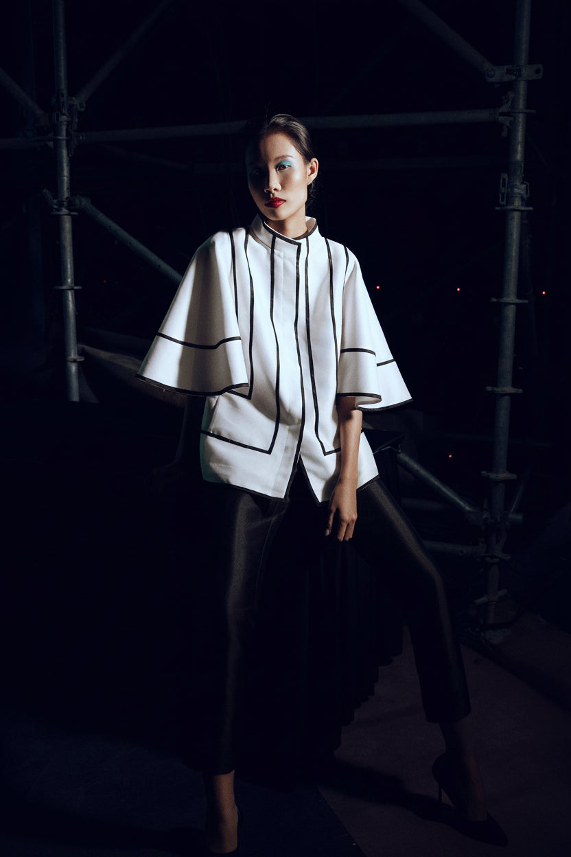 Model in a white cape-sleeved jacket edged in black, on a dark runway